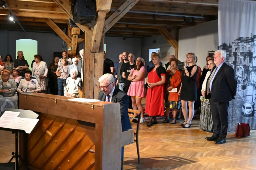 Uroczyste otwarcie wystawy w dużej sali z drewnianą konstrukcją i fortepianem na pierwszym planie; przy instrumencie siedzi mężczyzna, a za nim zgromadzona publiczność obserwuje wydarzenie; po prawej stronie stoją uczestnicy wernisażu, a ściany zdobią wielkoformatowe fotografie i materiały związane z wystawą (kliknięcie powiększy zdjęcie).
