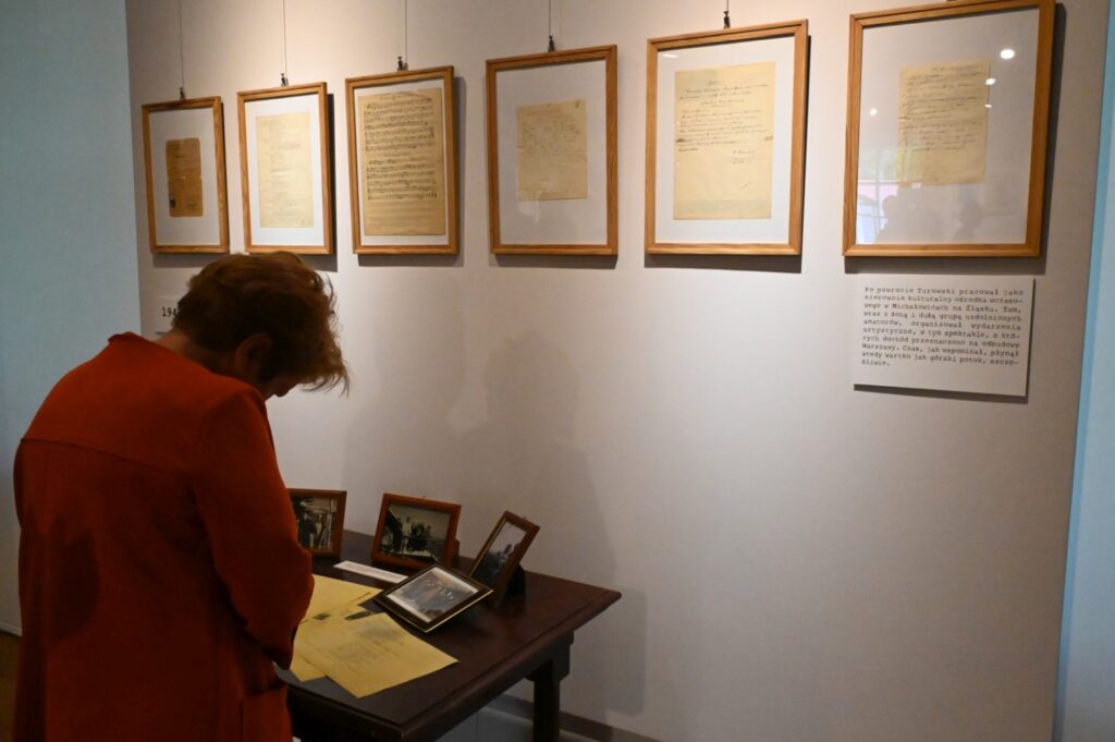 Fragment ekspozycji z szeregiem oprawionych dokumentów i archiwalnych materiałów zawieszonych na białej ścianie; poniżej znajduje się stół z fotografiami i dokumentami, przy którym pochyla się zwiedzająca osoba (kliknięcie powiększy zdjęcie).