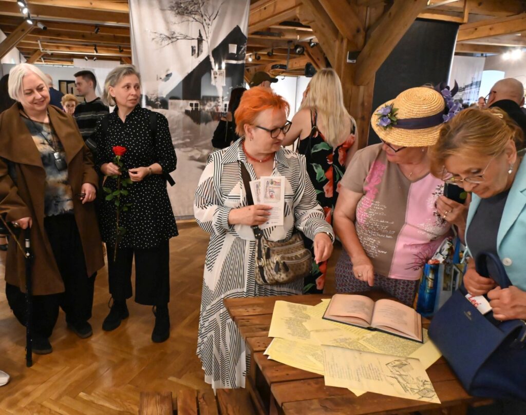 Zwiedzający oglądają dokumenty i pamiątki zgromadzone na stole; na pierwszym planie kobieta w jasnym stroju i kapeluszu pochyla się nad otwartym albumem, obok inne osoby przeglądają fotografie, dokumenty i katalogi; w tle widoczni są kolejni uczestnicy wydarzenia (kliknięcie powiększy zdjęcie).