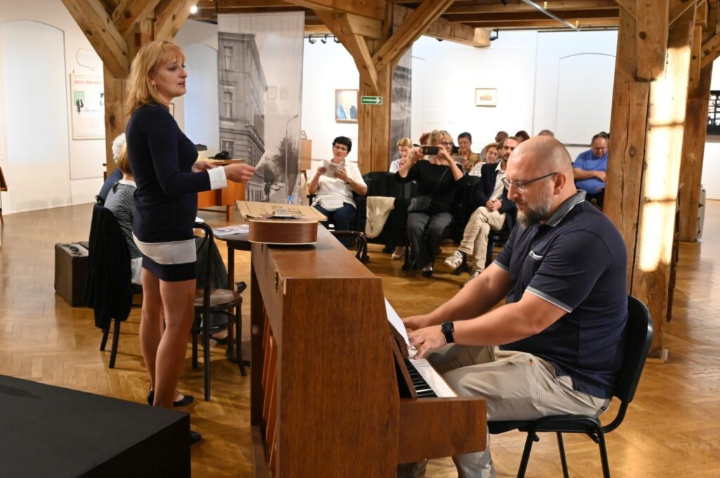 Kobieta stoi obok pianina i przemawia do zgromadzonych podczas wydarzenia muzyczno-literackiego (kliknięcie powiększy zdjęcie).