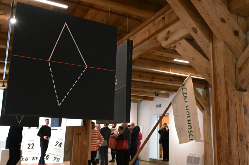 Fragment wystawy z geometryczną instalacją oraz widoczną przestrzenią galerii i zwiedzającymi w tle (kliknięcie powiększy zdjęcie).