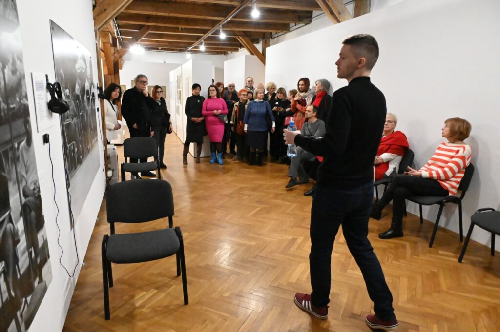 Przewodnik stoi przed grupą zwiedzających i omawia wystawę w sali z krzesłami ustawionymi pod ścianą (kliknięcie powiększy zdjęcie).