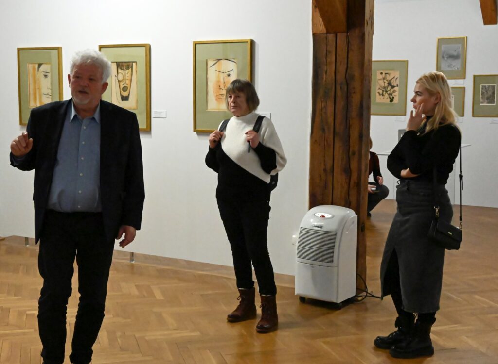 Mężczyzna oprowadza grupę po wystawie stojąc przy ścianie z pracami a uczestnicy słuchają jego wyjaśnień (kliknięcie powiększy zdjęcie).