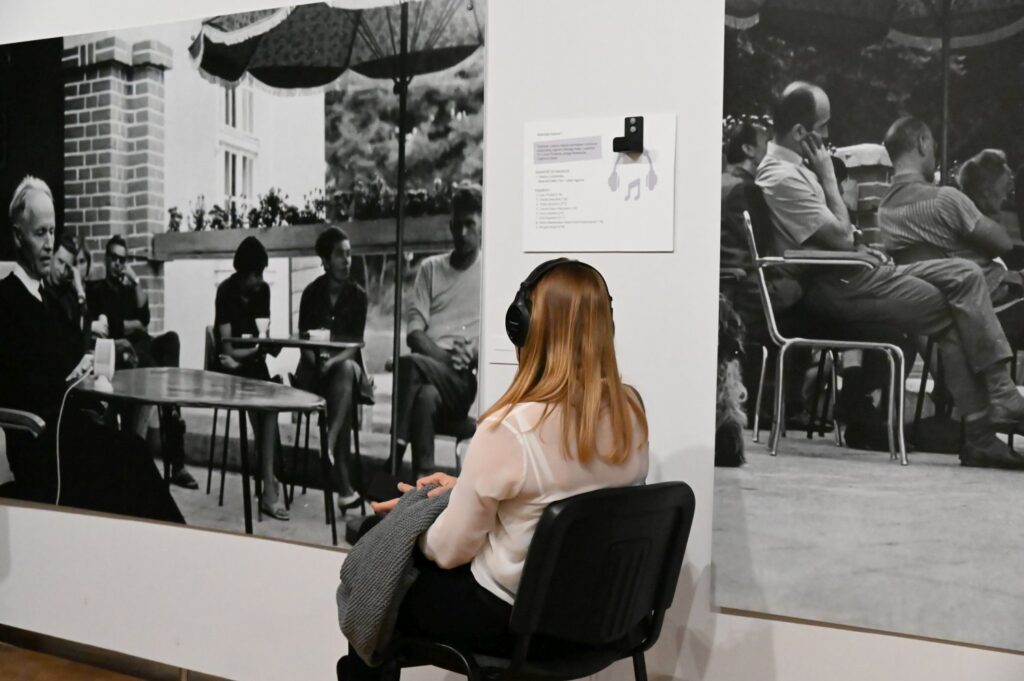 Kobieta siedzi na krześle i słucha nagrania przy stanowisku multimedialnym obok dużych fotografii na ścianie (kliknięcie powiększy zdjęcie).