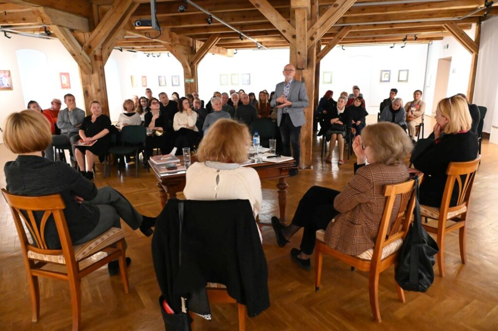 Widok sali wystawowej z uczestnikami siedzącymi w kręgu i słuchającymi wystąpienia w przestrzeni z drewnianymi belkami (kliknięcie powiększy zdjęcie).