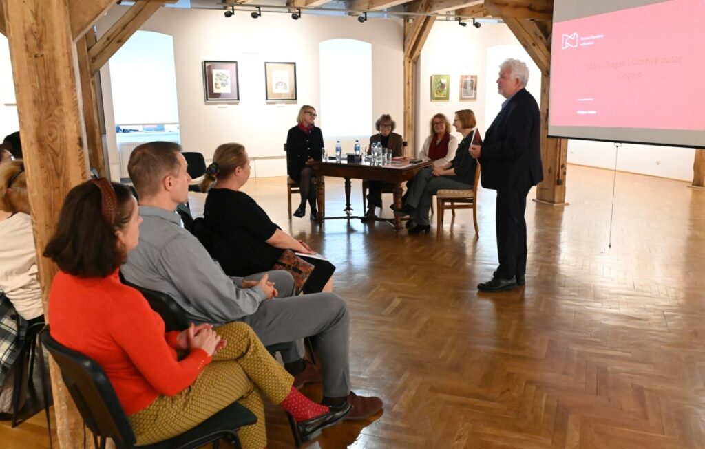 Uczestnicy konferencji siedzą na krzesłach w sali wystawowej a prowadzący przemawia stojąc przed nimi przy stole panelowym (kliknięcie powiększy zdjęcie).