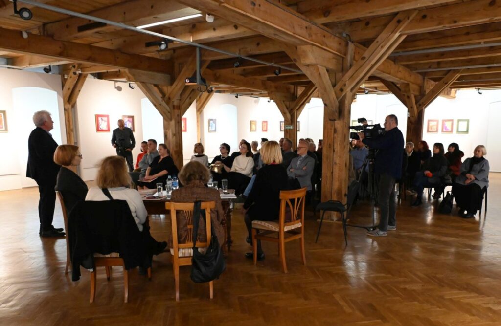 Prowadzący stoi na środku sali i przemawia do zgromadzonych uczestników konferencji (kliknięcie powiększy zdjęcie).