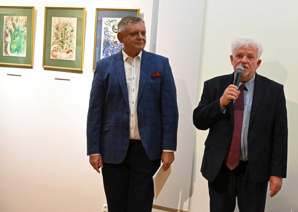 Dwaj mężczyźni podczas wernisażu stoją przed ścianą z kolorowymi grafikami; mężczyzna po prawej przemawia do zgromadzonych z mikrofonem, a drugi stoi obok niego z kartką w dłoni; w tle widoczne są trzy grafiki w zielonych i niebieskich ramach oraz fragment kolejnych prac należących do ekspozycji (kliknięcie powiększy zdjęcie).