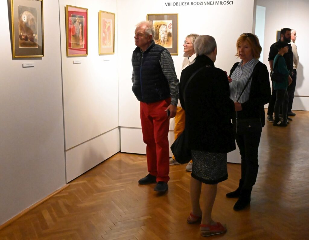 Przy ścianie stoi grupa kilku osób, które rozmawiają i przyglądają się grafikom; na pierwszym planie widoczny jest mężczyzna w czerwonych spodniach oraz dwie kobiety, a w głębi kolejne osoby zwiedzają wystawę (kliknięcie powiększy zdjęcie).