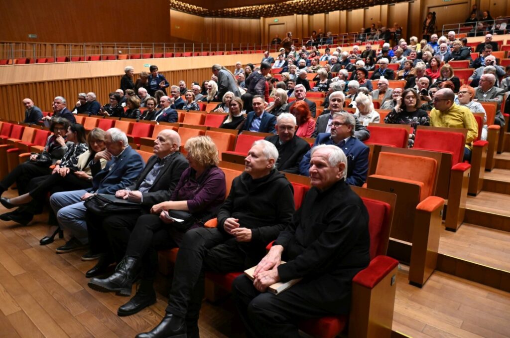 Publiczność siedząca w rzędach czerwonych foteli w sali koncertowej oczekuje na rozpoczęcie wydarzenia (kliknięcie powiększy zdjęcie).