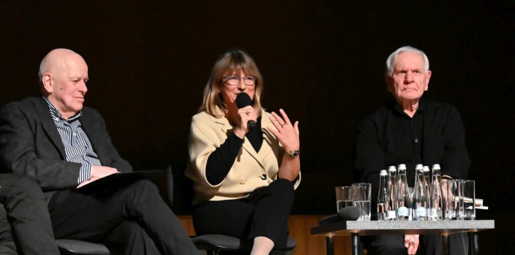 Kobieta siedząca na scenie trzyma mikrofon i zabiera głos podczas panelu dyskusyjnego obok inni uczestnicy (kliknięcie powiększy zdjęcie).
