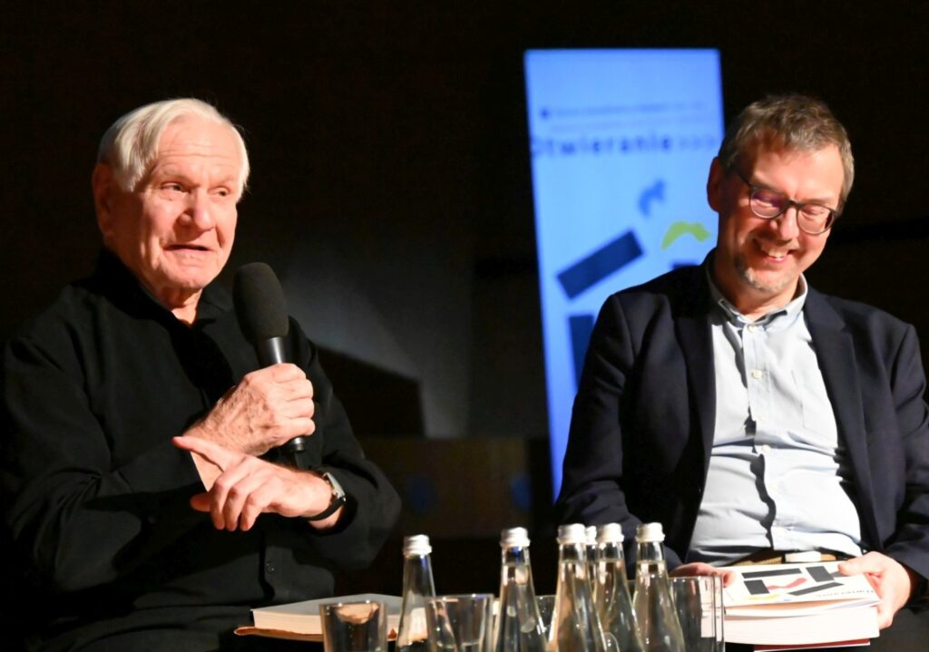 Starszy mężczyzna przemawia do mikrofonu siedząc na scenie obok inny panelista uśmiecha się trzymając materiały (kliknięcie powiększy zdjęcie).