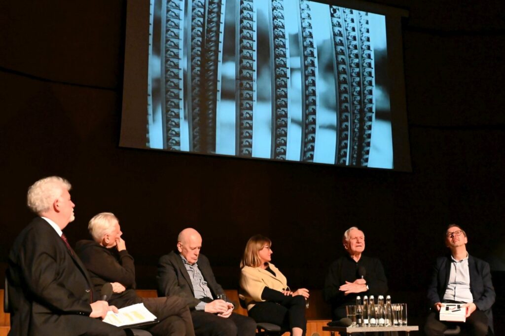 Paneliści siedzą na scenie i oglądają projekcję wyświetlaną na dużym ekranie podczas wydarzenia (kliknięcie powiększy zdjęcie).