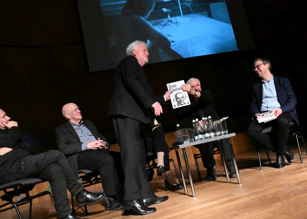Mężczyzna stojący na scenie pokazuje publikację panelistom siedzącym obok podczas spotkania (kliknięcie powiększy zdjęcie).