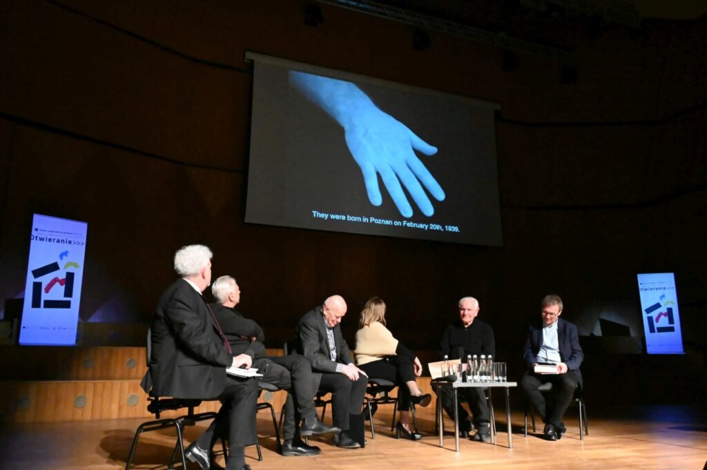 Paneliści siedzą na scenie i oglądają projekcję zbliżenia dłoni wyświetlaną na ekranie (kliknięcie powiększy zdjęcie).
