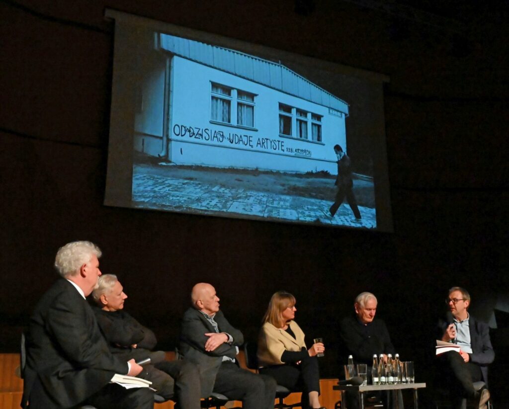 Panel dyskusyjny na scenie z projekcją budynku na ekranie w tle uczestnicy rozmawiają podczas wydarzenia (kliknięcie powiększy zdjęcie).