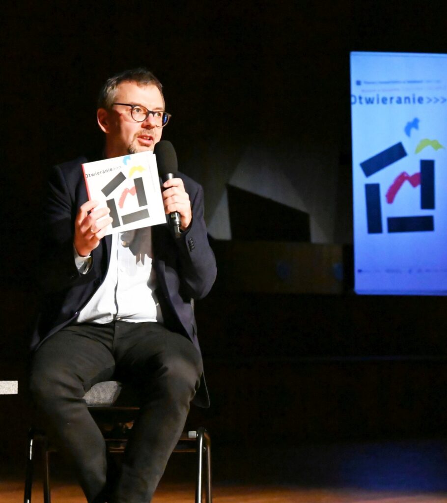 Mężczyzna siedzący na scenie trzyma mikrofon i pokazuje publikację podczas wydarzenia (kliknięcie powiększy zdjęcie).
