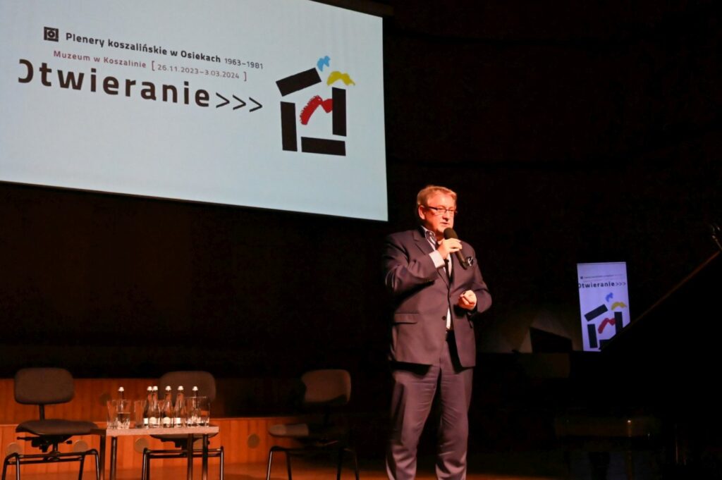 Prowadzący w garniturze przemawia na scenie na tle ekranu z napisem Otwieranie (kliknięcie powiększy zdjęcie).