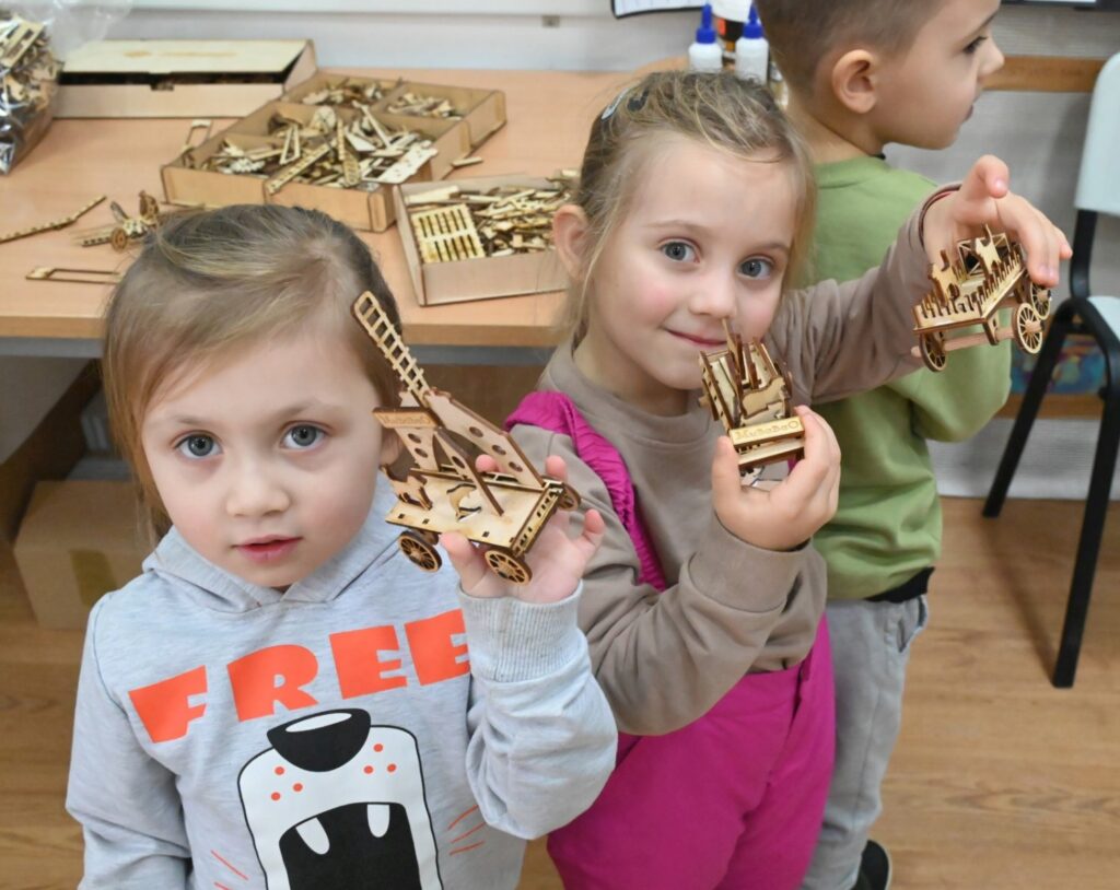 Dwie dziewczynki pokazują do kamery złożone drewniane modele, trzymając je w dłoniach (kliknięcie powiększy zdjęcie).