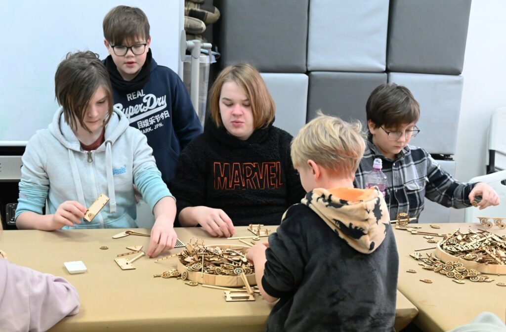Grupa dzieci siedzi przy stole i wspólnie wybiera oraz dopasowuje elementy drewnianych modeli (kliknięcie powiększy zdjęcie).