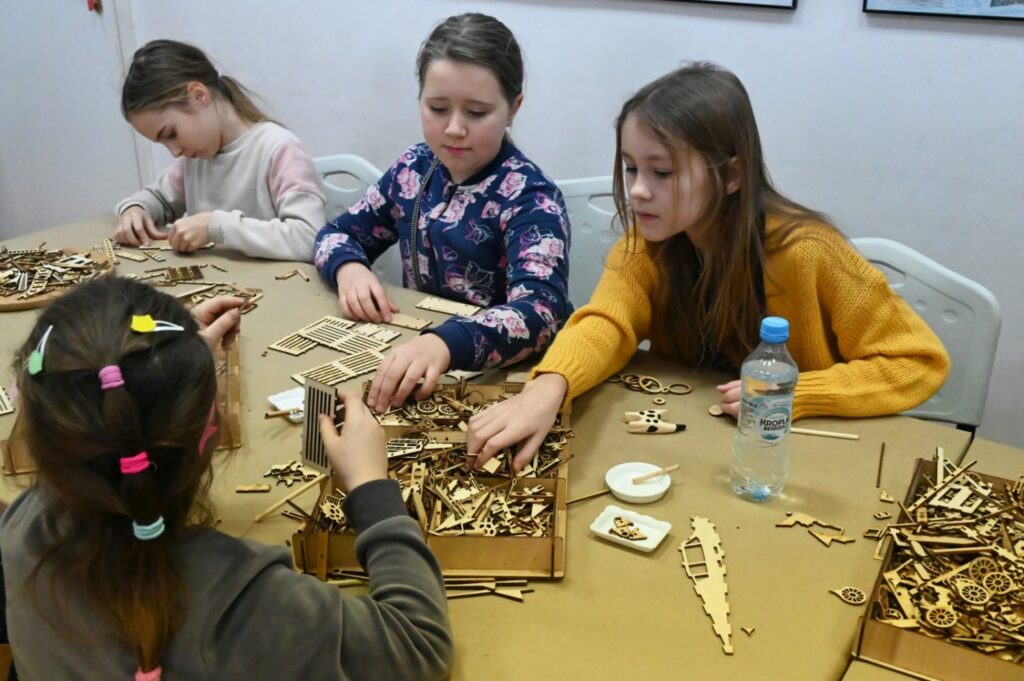 Kilkoro dzieci pracuje nad konstrukcjami z drewnianych części, układając elementy na stole pełnym materiałów (kliknięcie powiększy zdjęcie).