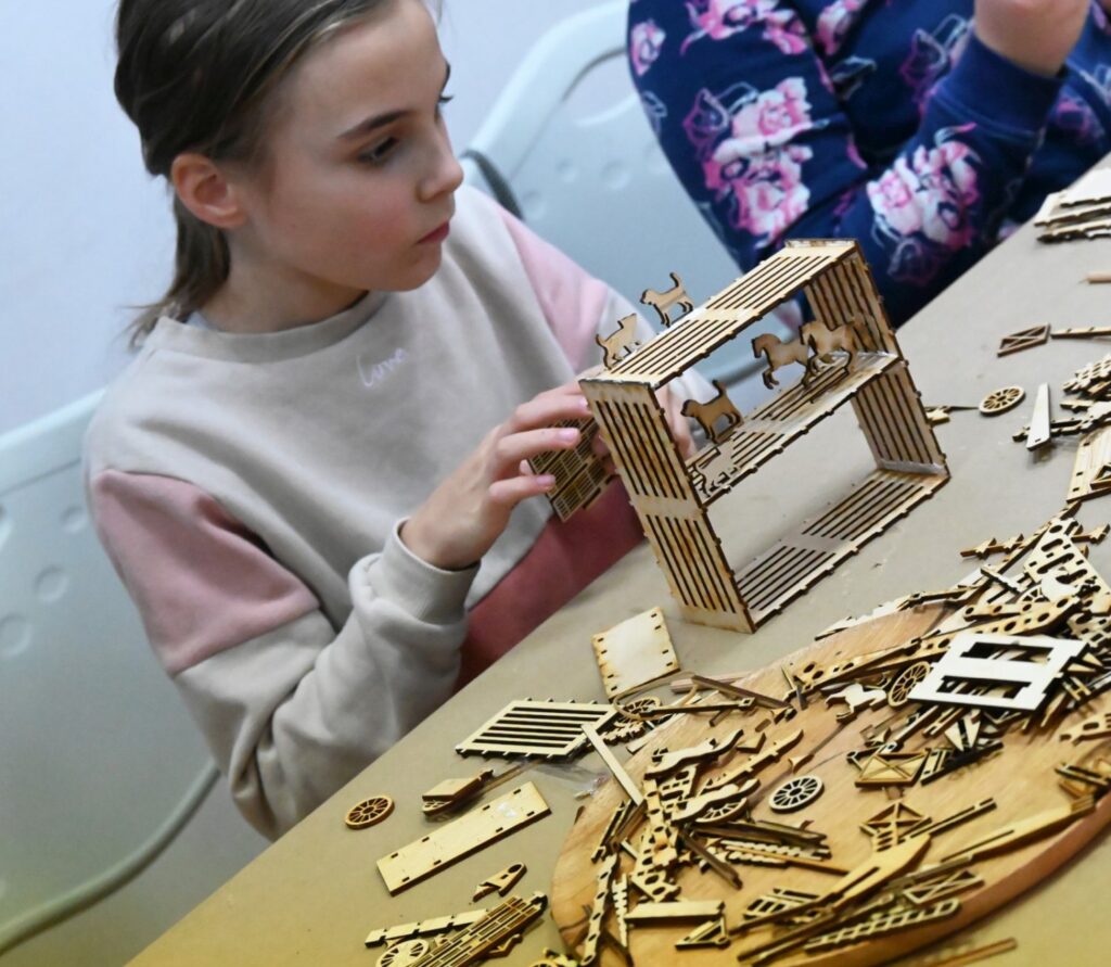 Dziewczynka uważnie przygląda się powstającej konstrukcji drewnianego modelu, trzymając go w dłoniach (kliknięcie powiększy zdjęcie).