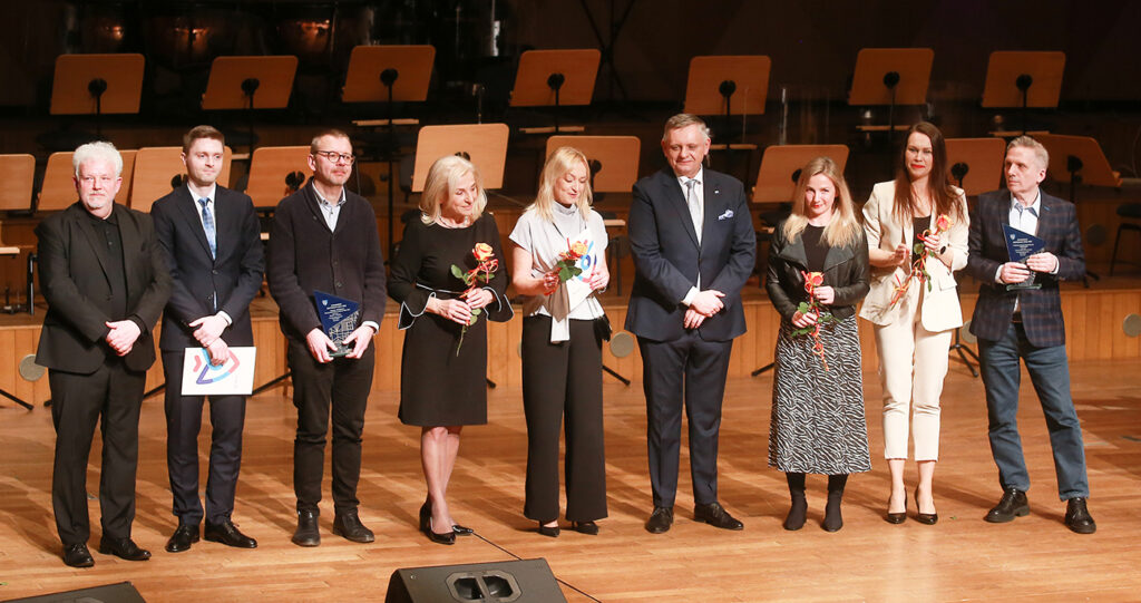 Grupa laureatów stojących na scenie sali koncertowej trzymających statuetki dyplomy i kwiaty w tle widoczne pulpity orkiestry (kliknięcie powiększy zdjęcie).