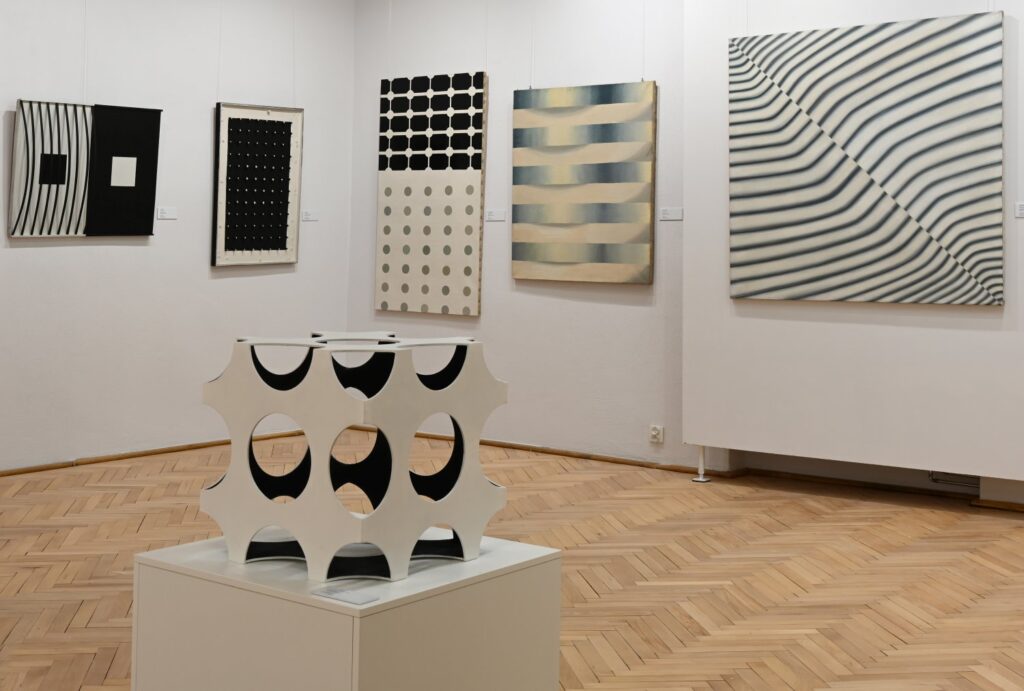Fragment wystawy z geometrycznymi kompozycjami na ścianach; na pierwszym planie stoi biała rzeźba o ażurowej formie z okrągłymi wycięciami, a w tle widoczne są obrazy z czarno-białymi wzorami i falistymi liniami (kliknięcie powiększy zdjęcie).