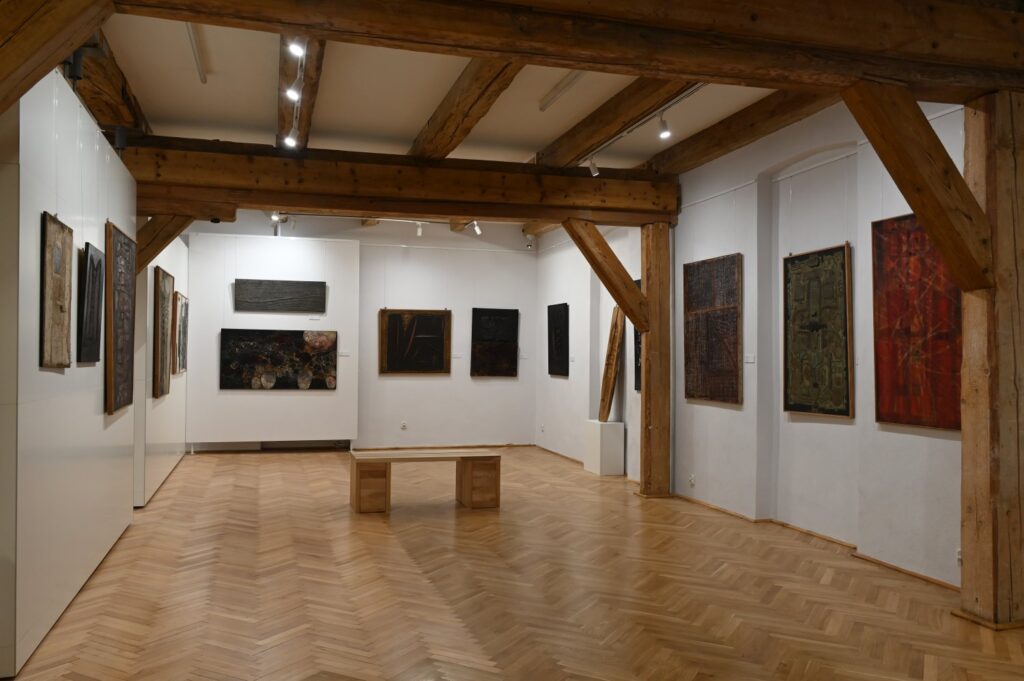 Sala wystawowa z obrazami sztuki współczesnej w różnych formatach i kolorach; prace rozmieszczono na białych ścianach, a na środku ustawiono drewnianą ławkę; wnętrze ma drewnianą konstrukcję stropu i parkietową podłogę (kliknięcie powiększy zdjęcie).