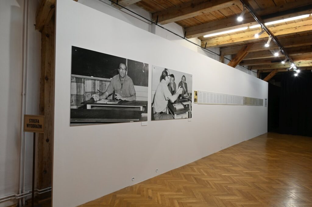 Fragment wystawy z czarno-białymi fotografiami dokumentalnymi; na ścianie widoczne są dwa duże zdjęcia przedstawiające osoby przy pracy oraz długi pas materiałów tekstowych; przed ekspozycją znajduje się wolne miejsce dla zwiedzających (kliknięcie powiększy zdjęcie).