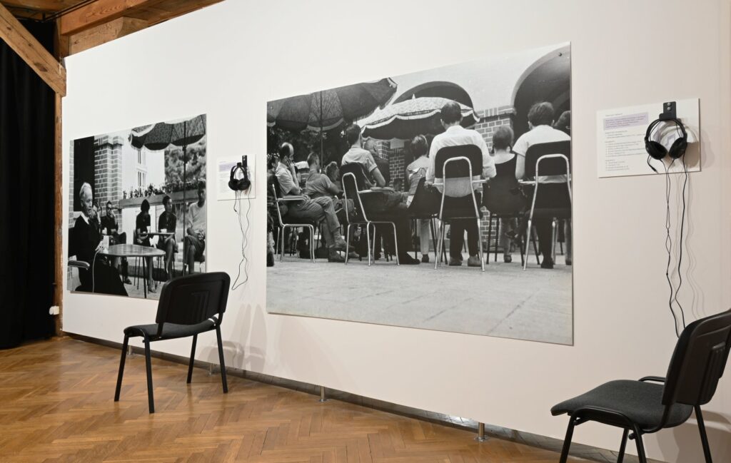 Fragment wystawy z dużymi czarno-białymi fotografiami; na ścianie widoczne są fotografie przedstawiające ludzi podczas spotkania oraz osoby siedzące przy stołach; przed ekspozycją ustawiono dwa krzesła i słuchawki do odsłuchu nagrań (kliknięcie powiększy zdjęcie).