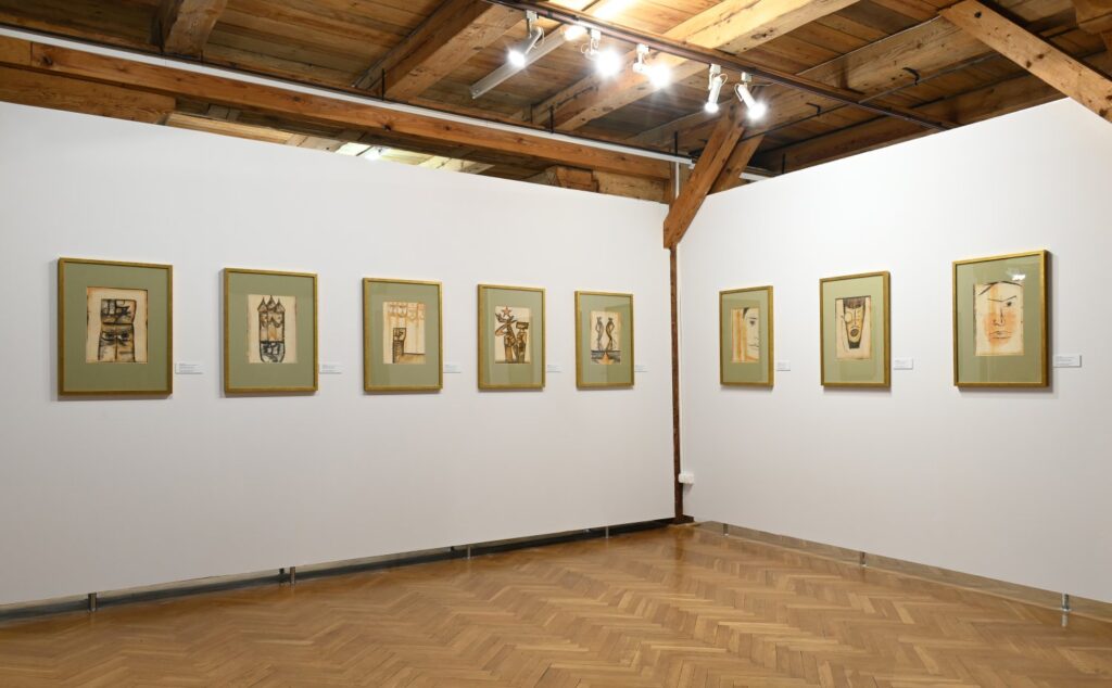 Sala wystawowa z pracami artystycznymi i dokumentacją; na prawej ścianie znajduje się seria niewielkich grafik i rysunków w złotych ramach, a na lewej duże czarno-białe fotografie; przed ekspozycją ustawiono krzesła dla zwiedzających (kliknięcie powiększy zdjęcie).