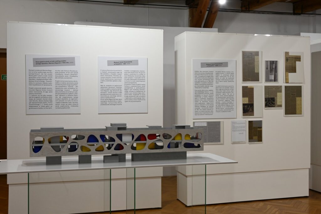 Fragment ekspozycji z tekstami oraz materiałami archiwalnymi; na białych ściankach znajdują się opisy, fotografie i reprodukcje dokumentów, a na pierwszym planie ustawiono podłużną metalową rzeźbę z kolorowymi elementami (kliknięcie powiększy zdjęcie).