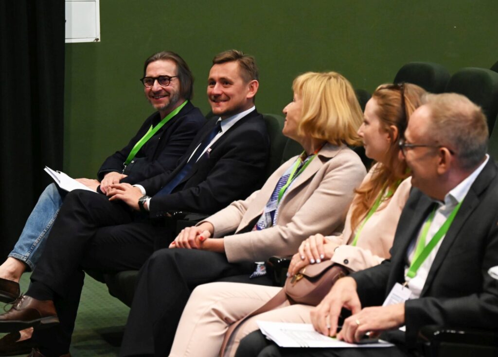 Goście siedzą w pierwszym rzędzie sali konferencyjnej, uśmiechając się i rozmawiając przed rozpoczęciem wydarzenia (kliknięcie powiększy zdjęcie).