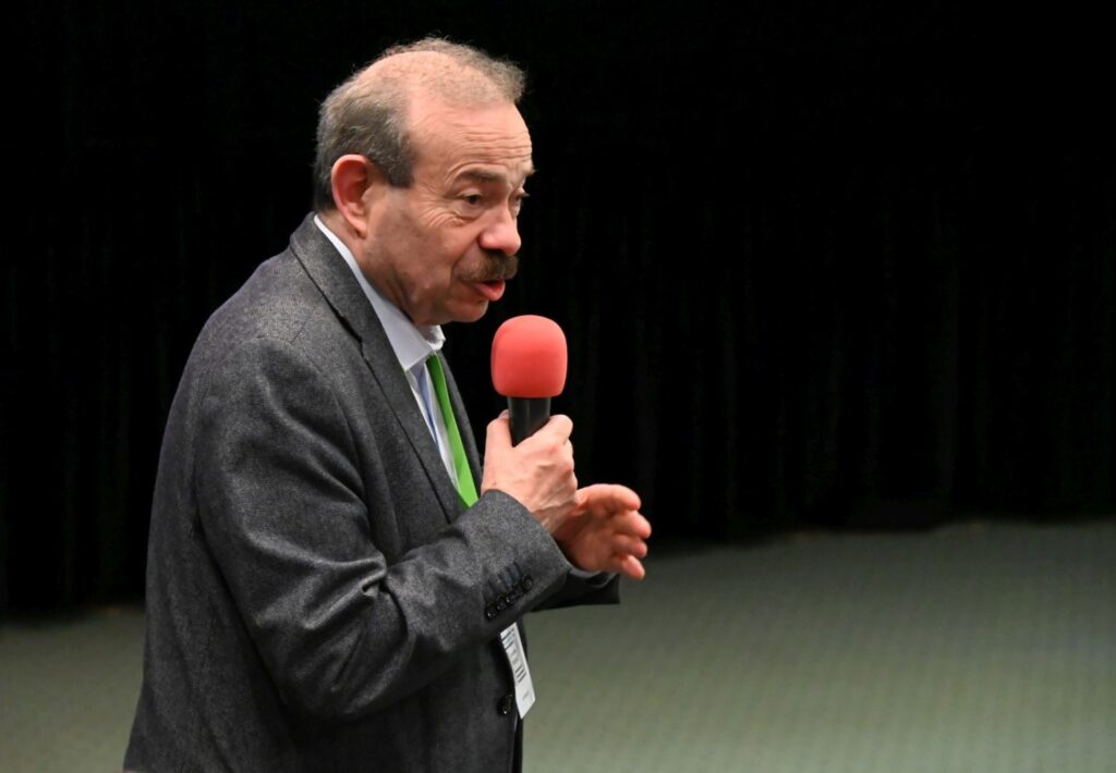 Prelegent trzyma mikrofon i zwraca się do uczestników konferencji podczas wystąpienia (kliknięcie powiększy zdjęcie).