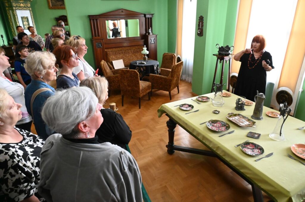 Przewodniczka na sali wystawy sztuki dawnej opowiada o eksponatach przy stole, a uczestnicy słuchają w zabytkowym pomieszczeniu muzealnym (kliknięcie powiększy zdjęcie).