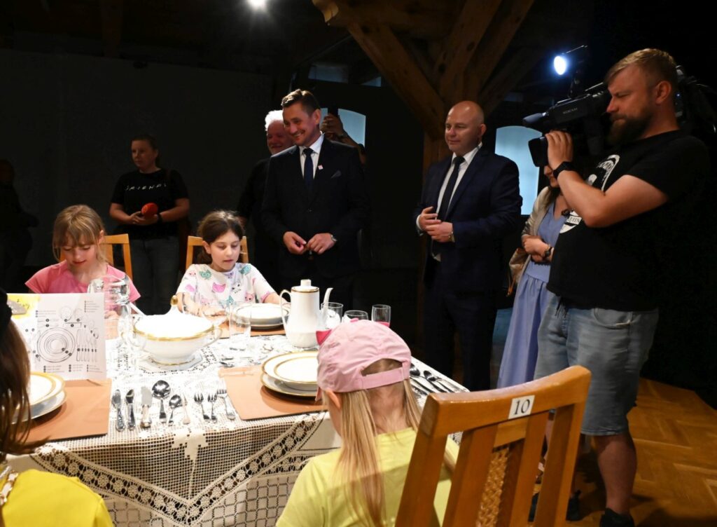 Dzieci siedzą przy stole podczas warsztatów, a wokół stoją dorośli i operator kamery nagrywający wydarzenie (kliknięcie powiększy zdjęcie).