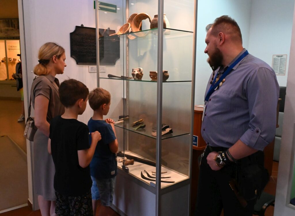 Kobieta z dwójką dzieci ogląda zabytki w szklanej gablocie, a pracownik muzeum stoi obok i obserwuje (kliknięcie powiększy zdjęcie)..