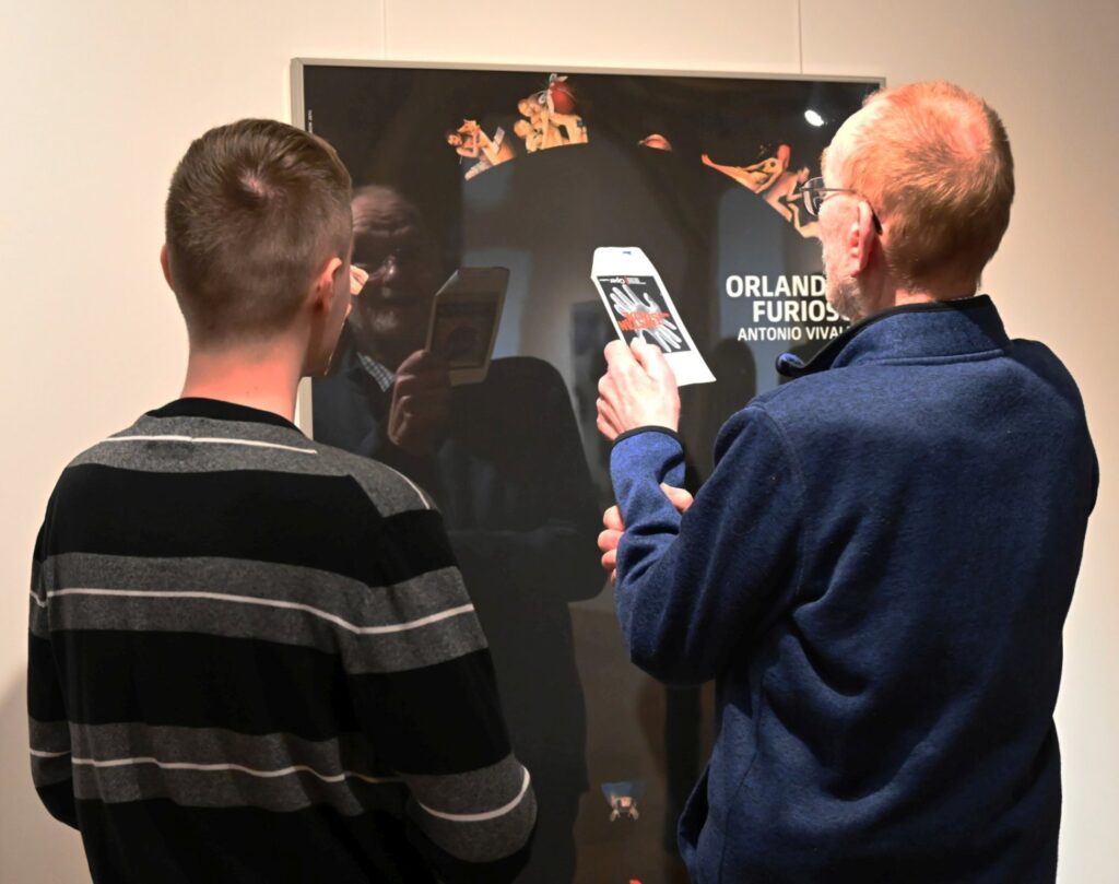 Dwóch uczestników ogląda plakat przedstawiający spektakl „Orlando furioso”; starszy mężczyzna trzyma w dłoni ulotkę o wystawie (kliknięcie powiększy zdjęcie).