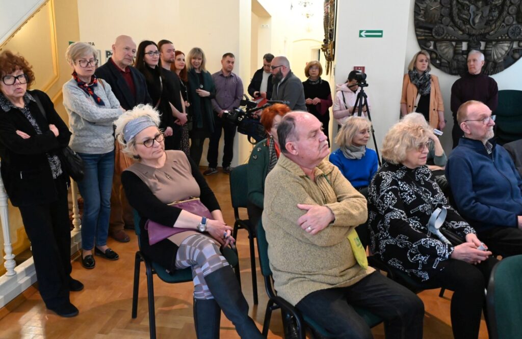 Uczestnicy wydarzenia zgromadzeni w muzealnej sali podczas oficjalnej części spotkania; część osób siedzi na krzesłach, pozostali stoją wzdłuż ściany i przy wejściu do sali; w tle widoczne są schody oraz elementy architektury zabytkowego wnętrza; fotografia pokazuje liczną publiczność uczestniczącą w otwarciu wystawy (kliknięcie powiększy zdjęcie).