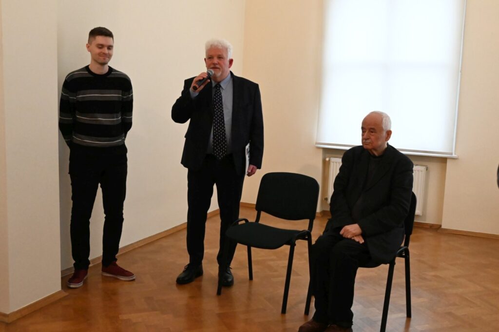 Mężczyzna stojący przy mikrofonie zwraca się do zgromadzonych gości; obok niego stoi młody mężczyzna, a na krześle siedzi starszy uczestnik wydarzenia; w tle widoczna jest jasna sala muzealna z dużym oknem oraz fragment ekspozycji; fotografia dokumentuje oficjalną część wernisażu (kliknięcie powiększy zdjęcie).