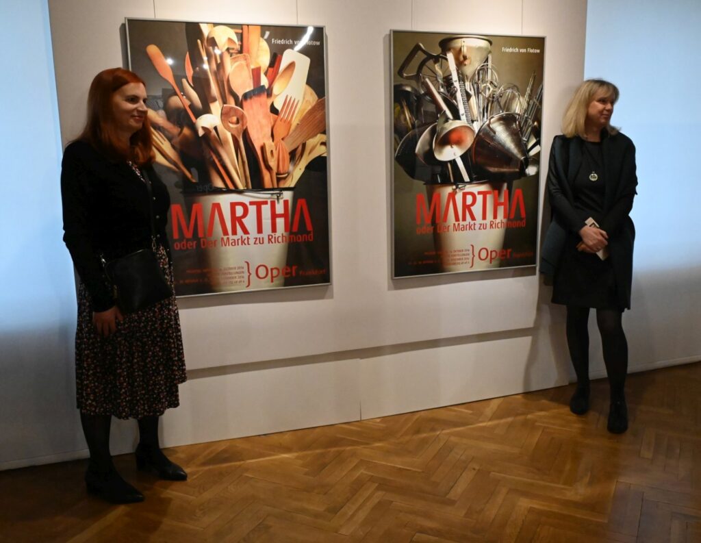 Dwie kobiety pozują przy dwóch plakatach przedstawiających operę „Martha oder Der Markt zu Richmond”; jedna z kobiet stoi bliżej pierwszego planu, druga znajduje się przy ścianie ekspozycyjnej (kliknięcie powiększy zdjęcie).