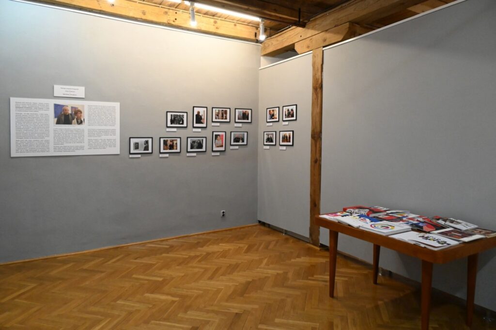 Widok ściany z materiałami dokumentacyjnymi i fotografiami; ekspozycja obejmuje teksty informacyjne, reprodukcje oraz archiwalne fotografie przedstawione w niewielkich ramach; w sali znajduje się również stół z publikacjami (kliknięcie powiększy zdjęcie).