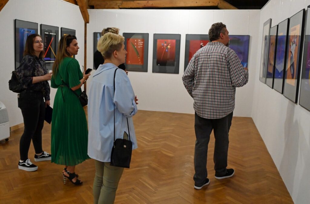 Uczestnicy wernisażu oglądają kolorowe prace graficzne; na pierwszym planie znajduje się grupa zwiedzających, a na ścianach widoczne są abstrakcyjne kompozycje w intensywnych barwach (kliknięcie powiększy zdjęcie).