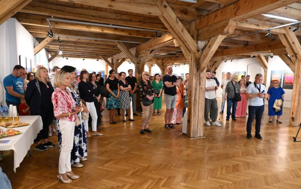 Szerszy widok wernisażu; liczna grupa uczestników zgromadziła się w sali wystawowej, część osób ogląda prace, a część uczestniczy w spotkaniu; w tle widoczne są kolejne części ekspozycji (kliknięcie powiększy zdjęcie).