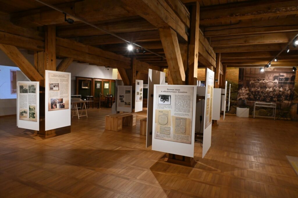 Widok sali wystawowej z ekspozycją poświęconą historii miasta i lokalnego przemysłu; na drewnianych stojakach ustawiono plansze z fotografiami, dokumentami i tekstami, w głębi widoczna jest duża archiwalna fotografia (kliknięcie powiększy zdjęcie).