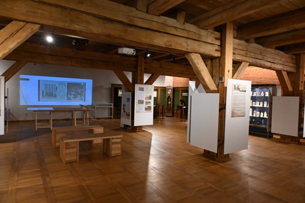 Widok sali wystawowej z planszami informacyjnymi, gablotami i ekranem prezentującym materiały multimedialne; wnętrze ma drewnianą konstrukcję i parkietową podłogę (kliknięcie powiększy zdjęcie).