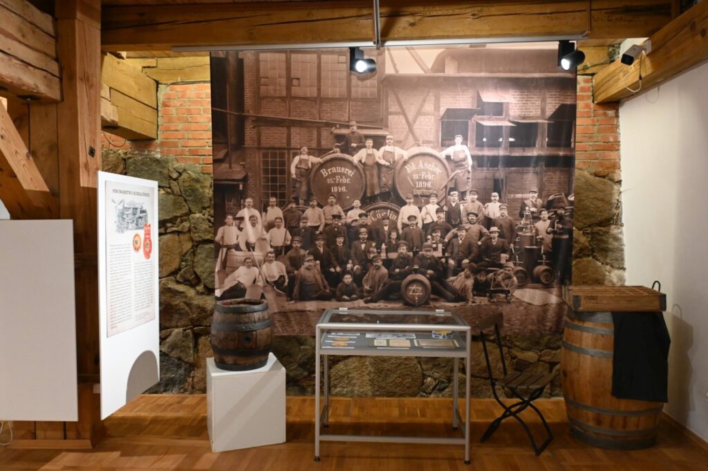 Archiwalna fotografia przedstawiająca grupę pracowników browaru; przed wielkoformatowym zdjęciem ustawiono beczki, gablotę z eksponatami oraz planszę informacyjną (kliknięcie powiększy zdjęcie).