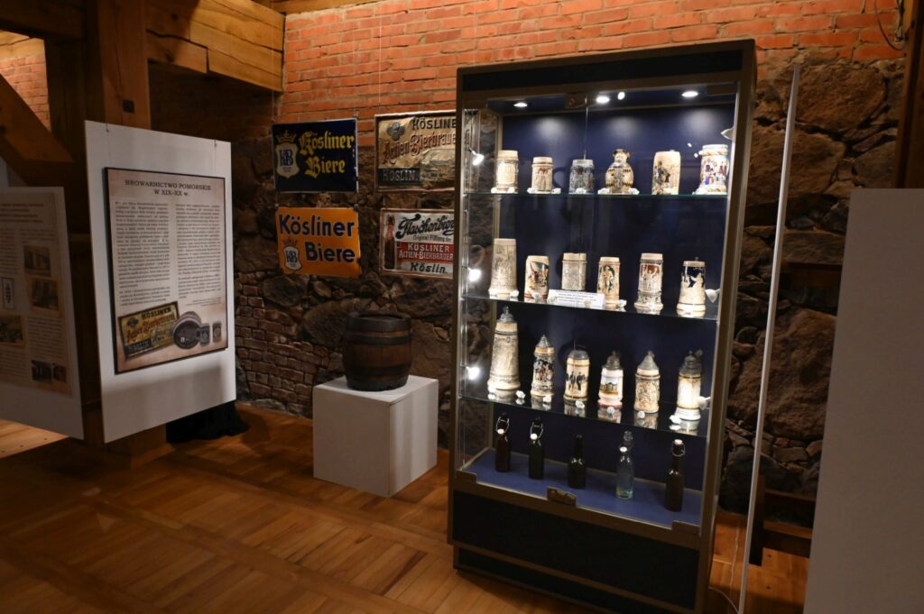 Gablota z kolekcją historycznych butelek i naczyń; obok znajduje się plansza informacyjna oraz reklamy piwa, a ekspozycję umieszczono na tle ceglanej i kamiennej ściany (kliknięcie powiększy zdjęcie).