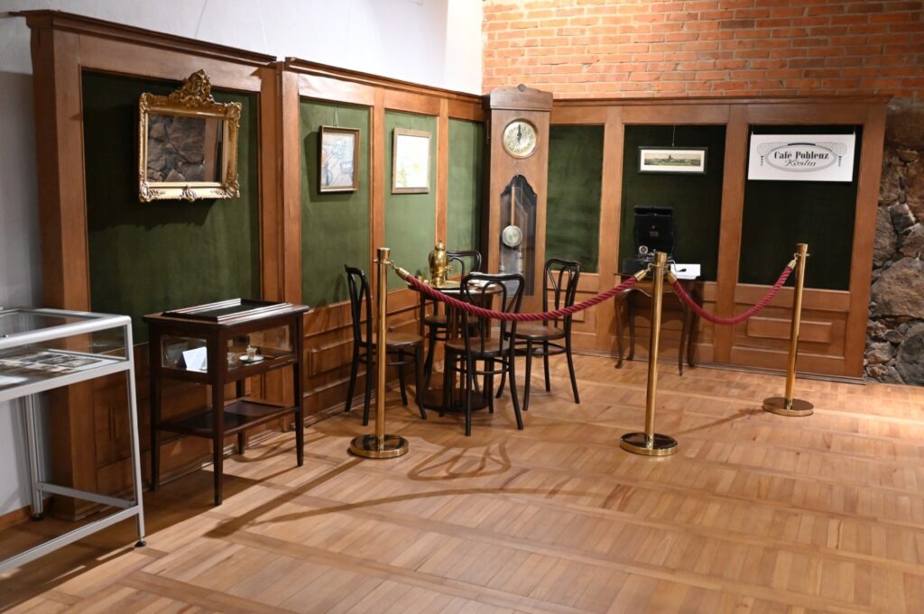 Fragment wystawy zaaranżowany jako historyczne wnętrze; przy ścianie ustawiono stoliki i krzesła, zegar oraz zabytkowe przedmioty, a ekspozycję odgrodzono czerwonymi linami na słupkach (kliknięcie powiększy zdjęcie).